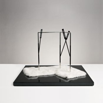 A minimalist sculpture with a metal geometric frame stands on a glossy black base, which is partly covered with two irregularly shaped pieces of white fur. The background is gray and uncluttered.